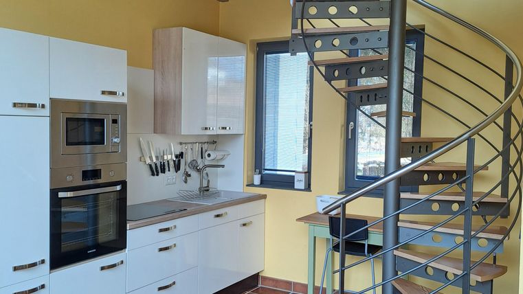 Eine moderne Küche mit weißen Schränken und einer geschwungenen Treppe. Die Wände sind in einem warmen Gelb gehalten und der Boden ist gefliest.