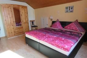 Ein gemütliches Schlafzimmer mit einem großen Bett und pinken Kissen. Holzschränke und ein Stuhl runden die Einrichtung ab.
