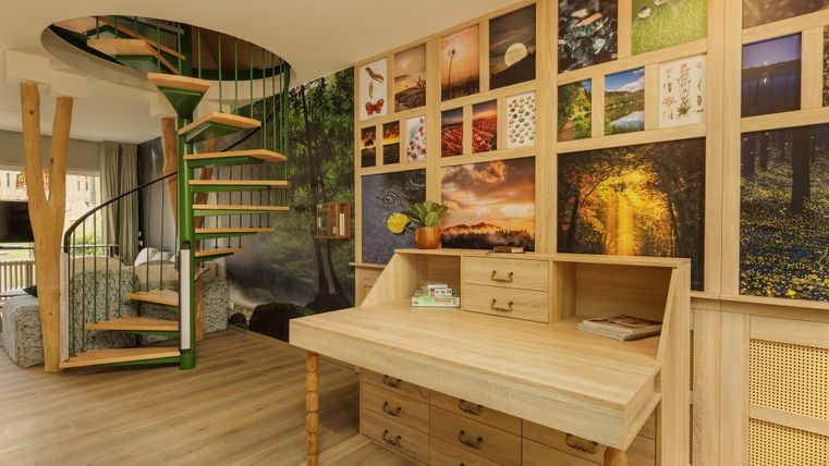 Ein moderner Wohnraum mit einer Holztreppe und einer Fülle von bunten Bildern an der Wand. Ein Holztisch mit Schubladen steht im Vordergrund, umgeben von einem hellen, einladenden Ambiente.