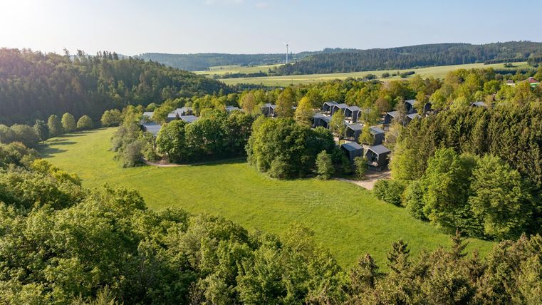 Eine ruhige Landschaft mit grünen Wiesen und Bäumen. Im Hintergrund sind Ferienhäuser und sanfte Hügel sichtbar.