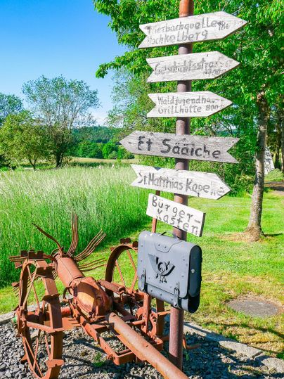 Ein rustikales Schild mit mehreren Wegweisern steht neben einem alten Traktor in der Natur. Im Hintergrund sind Bäume und grüne Wiesen zu sehen.