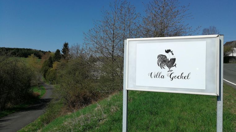 Ein Schild mit der Aufschrift "Villa Gochel" steht am Straßenrand. Im Hintergrund sind Bäume und ein klarer blauer Himmel zu sehen.