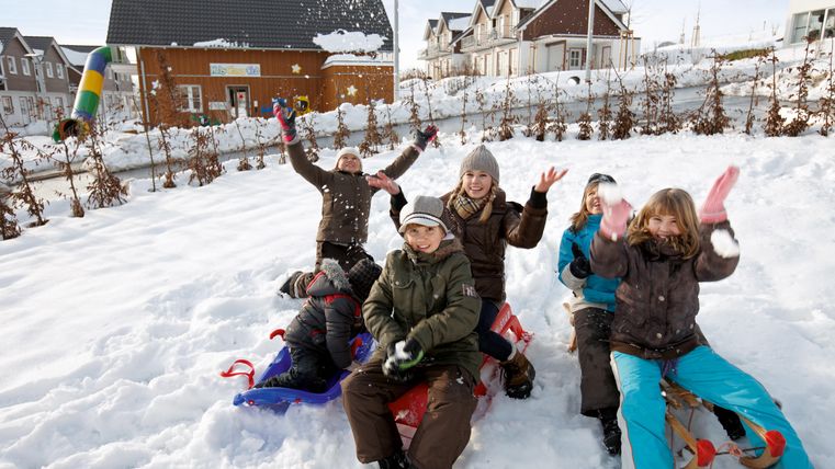Eine Gruppe von Kindern spielt fröhlich im Schnee und hat Schlitten dabei. Im Hintergrund sind schneebedeckte Häuser zu sehen.