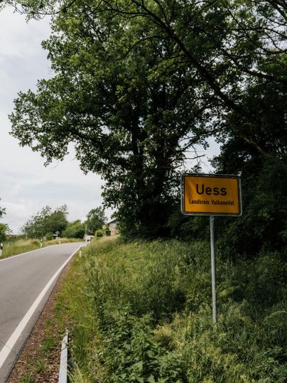 Ein Straßenschild mit der Aufschrift "Uess" steht am Rand einer kurvenreichen Landstraße. Umgeben von Bäumen und Grünflächen vermittelt die Szene eine ruhige ländliche Atmosphäre.