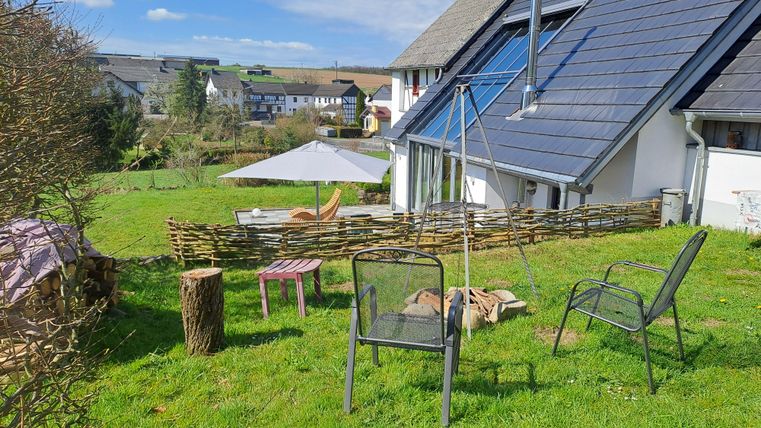 Ein gemütlicher Garten mit Rasen, mehreren Stühlen und einem Tisch. Im Hintergrund ist ein ruhiges Wohnhaus und ein Sonnenschirm zu sehen.