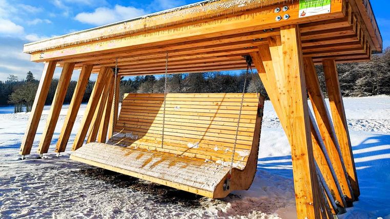 Eine große Holzschaukel steht im Schnee unter einem überdachten Rahmen. Der Himmel ist klar und blau.