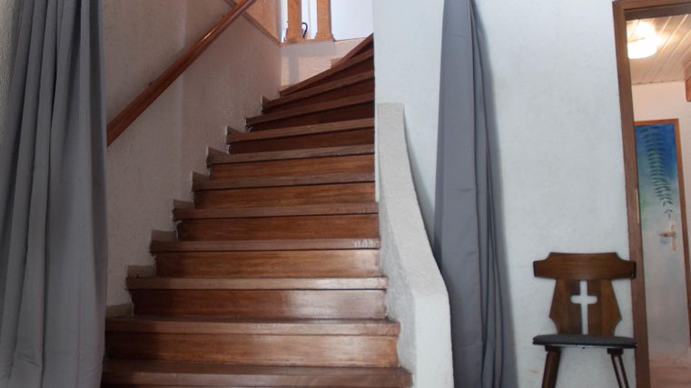 Eine Holztreppe führt nach oben, umgeben von grauen Vorhängen. Im Raum steht ein Stuhl mit einem Kreuz.