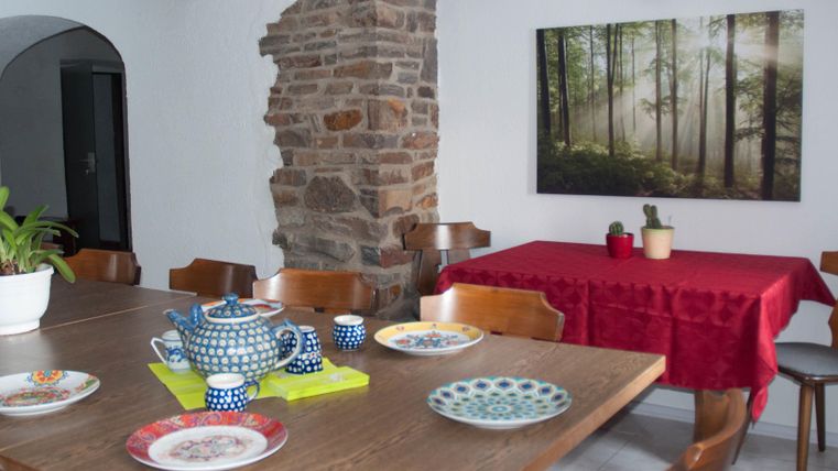 Ein einladender Essbereich mit einem großen Holztisch und Stühlen. Auf dem Tisch stehen bunte Teller und eine Teekanne, während ein Naturbild an der Wand hängt.