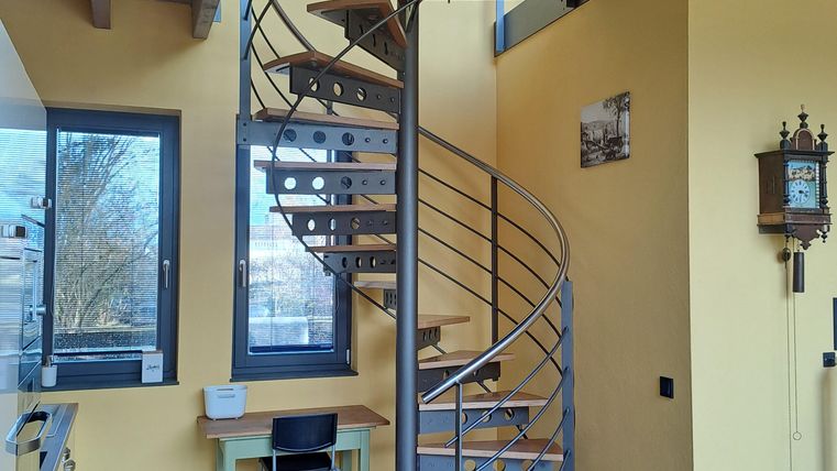 Eine moderne Wendeltreppe in einem hellen Raum mit gelben Wänden. Der Boden ist mit braunen Fliesen ausgelegt, und es gibt einen kleinen Tisch mit einem Stuhl.