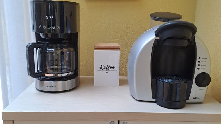 Eine Kaffeemaschine und eine Kaffeekapselmaschine stehen auf einem modernen Sideboard. Im Hintergrund hängt ein gerahmtes Bild mit einer Pflanzenzeichnung.