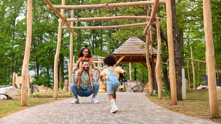 Ein fröhlicher Vater mit zwei Kindern spielt im Park. Die Kinder lachen und rennen auf einem gepflasterten Weg zwischen Bäumen und Spielgeräten.