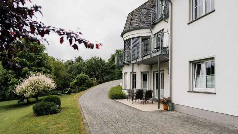 Ein modernes Haus mit einer großen Terrasse und einem gepflasterten Weg. Der Garten ist grün und gut gepflegt, umgeben von Bäumen und Sträuchern.