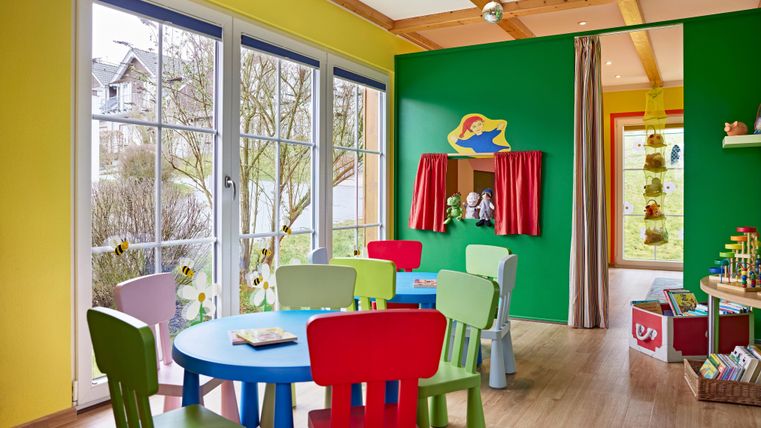 Ein bunter Spielraum mit einem runden Tisch und mehreren farbigen Stühlen. Die Wände sind in fröhlichen Farben gestrichen, und große Fenster lassen viel Licht herein.
