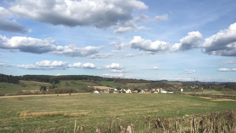 Ein weitläufiges Feld mit sanften Hügeln und einem kleinen Dorf im Hintergrund. Der Himmel ist blau mit vielen weißen Wolken.
