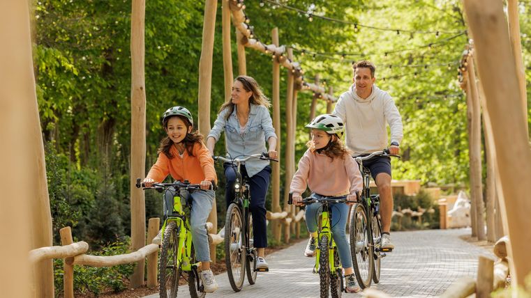 Eine Familie fährt mit Fahrrädern auf einem schmalen Weg durch einen grünen Park. Die Kinder tragen Helme und lächeln, während sie die Zeit im Freien genießen.