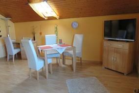 Ein gemütlicher Essbereich mit einem Tisch und weißen Stühlen. Im Raum befinden sich ein Fernseher und ein schöner Holzfußboden.