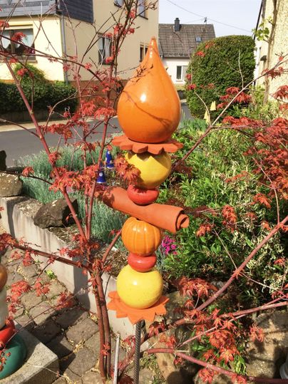 Ein bunter Gartendeko-Pfosten mit verschiedenen Formen in orange und gelb. Im Hintergrund sind herbstliche Äste und ein ruhiger Straßenblick zu sehen.