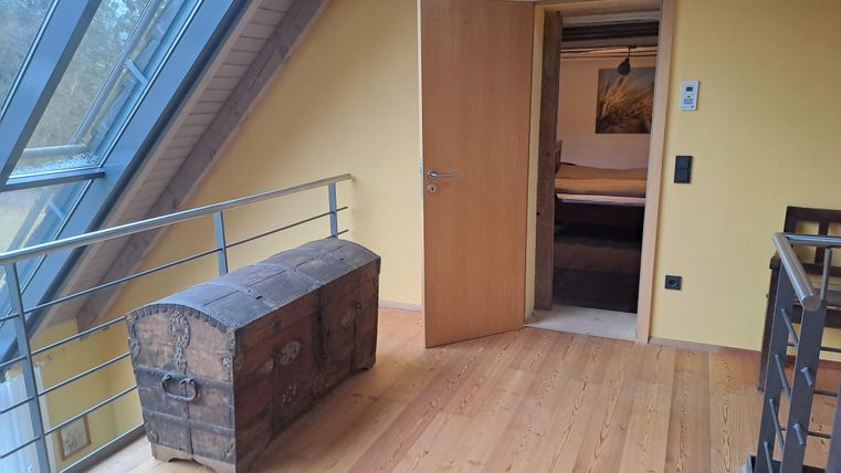 Ein heller Raum mit Holzboden und großen Fenstern. Eine offene Tür führt zu einem abgedunkelten Bereich, während eine antike Truhe im Vordergrund steht.
