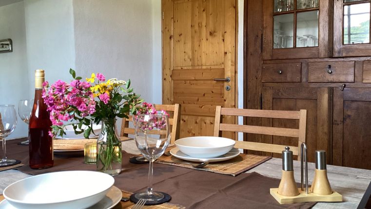 Ein schön gedeckter Tisch mit weißen Tellern und einer Blumenvase. Im Hintergrund sieht man eine Holzkommode und eine offene Tür.
