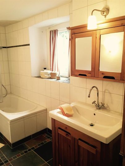 Ein helles Badezimmer mit einer Badewanne und einem Waschbecken aus Keramik. Holzelemente und Fenster mit Vorhängen schaffen eine einladende Atmosphäre.