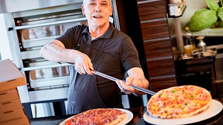 Ein älterer Mann in einem Restaurant schneidet gerade eine Pizza. Im Hintergrund sind weitere Pizzen und eine moderne Küche zu sehen.