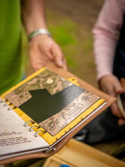 Ein offenes Notizbuch mit einer Karte und einem grünen Bereich ist zu sehen. Eine Hand hält das Buch, während eine andere Person darauf schaut.