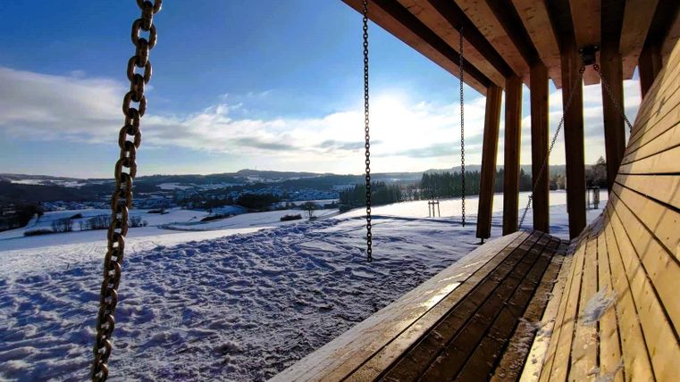 Eine große Holzschaukel steht in einer verschneiten Landschaft. Der Himmel ist klar und die Berge sind im Hintergrund sichtbar.