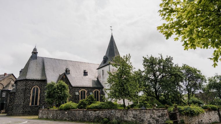 Eine malerische Kirche umgeben von Bäumen und Pflanzen. Der Himmel ist bewölkt, was eine ruhige Atmosphäre schafft.