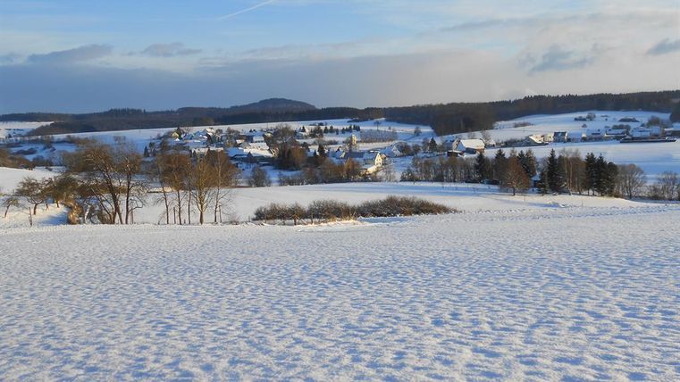 Eine verschneite Landschaft mit sanften Hügeln und verstreuten Häusern. Der Himmel ist klar und zeigt einen blauen Farbton.