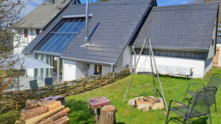 Ein modernes Haus mit großen Fenstern und einem schrägen Dach. Im Vordergrund sieht man eine Rasenfläche mit Holzstapeln und Gartenmöbeln.