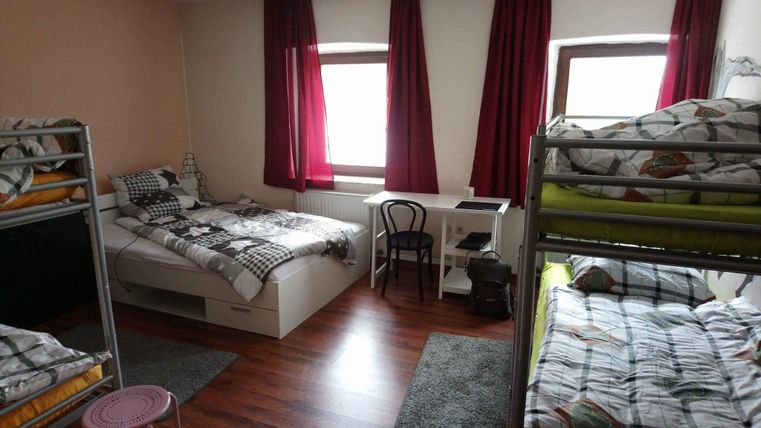 Ein einfaches Zimmer mit zwei Etagenbetten und einem Doppelbett. Tageslicht fällt durch die Fenster, und es gibt einen Schreibtisch mit einem Stuhl.
