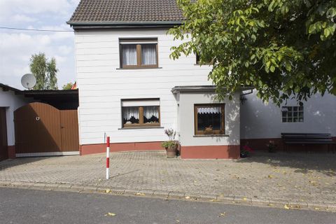 Ein Einfamilienhaus mit weißer Fassade und braunen Fensterrahmen. Vor dem Haus gibt es eine gepflasterte Fläche und einige Pflanzen.