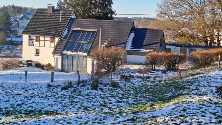 Ein modernes Haus mit großen Fenstern steht in einer verschneiten Landschaft. Im Hintergrund sind Bäume und ein klarer Himmel sichtbar.