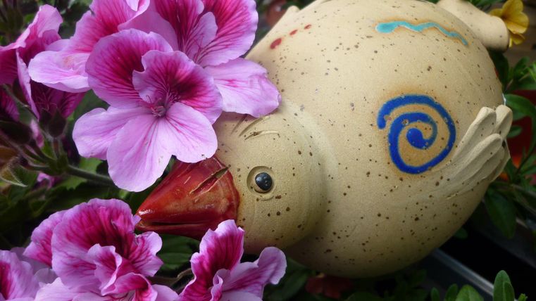 Ein buntes, dekoratives Huhn aus Keramik liegt zwischen lila Blumen. Die Blumen umrahmen das Huhn und verleihen der Szene eine fröhliche Atmosphäre.