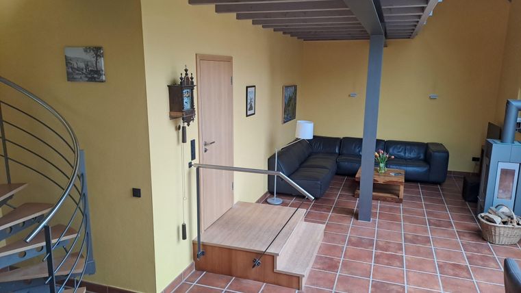 Ein heller Wohnraum mit gelben Wänden und einem modernen, grauen Sofa. An der Wand hängen Bilder, und eine Treppe führt in die obere Etage.