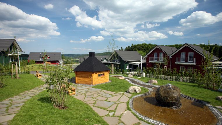 Eine malerische Landschaft mit mehreren bunten Holzhäusern. Der Garten ist grün und gepflegt, mit einem Teich und einer Feuerstelle in der Mitte.
