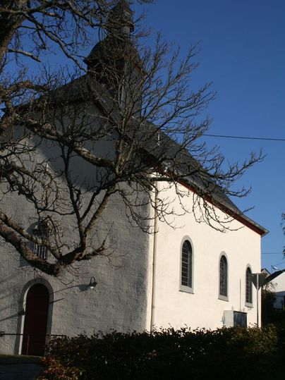 Eine weiße Kapelle umgeben von Bäumen und Sträuchern. Der Himmel ist klar und blau.