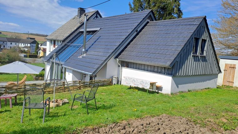 Ein modernes Haus mit einem gepflasterten Dach und großen Fenstern. Der Garten ist grün mit einigen Gartenmöbeln und Blick auf die umliegende Landschaft.