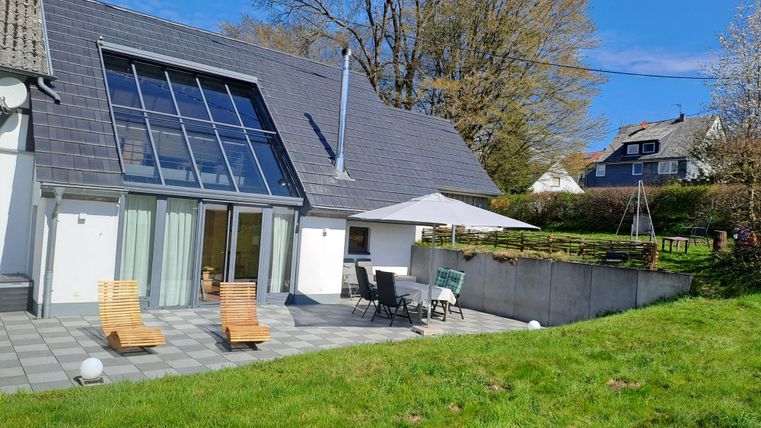 Ein modernes Haus mit großen Fenstern und einer Terrasse. Der Garten ist grün und einladend, ideal für entspannte Tage im Freien.