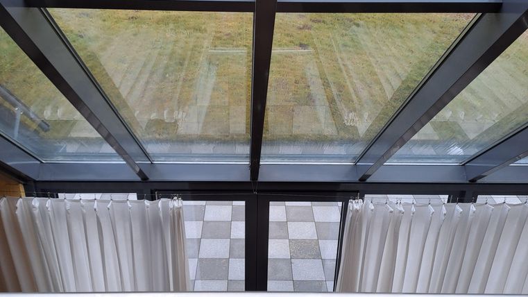 Ein moderner Raum mit großen Fenstern und blick auf eine grüne Landschaft. Vor den Fenstern hängen helle Vorhänge.