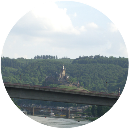 Eine malerische Landschaft mit einem Schloss auf einem grünen Hügel. Im Vordergrund verläuft eine Brücke über einen Fluss.
