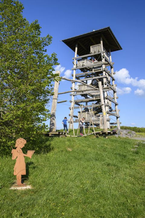 Ein Aussichtsturm aus Holz steht auf einer Wiese. Im Vordergrund ist eine metallene Figur, die ein Buch hält.