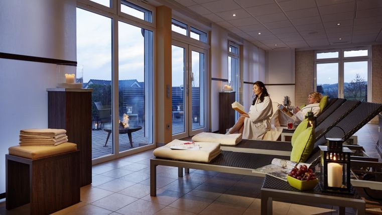 Ein ruhiger Spa-Bereich mit bequemen Liegen und großen Fenstern. Zwei Frauen entspannen sich, eine davon liest ein Buch.