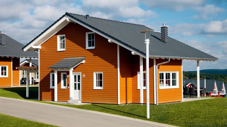 Ein modernes, orangefarbenes Haus mit einem grauen Dach. Das Gebäude steht auf einer grünen Wiese und ist von einer ruhigen Straße umgeben.