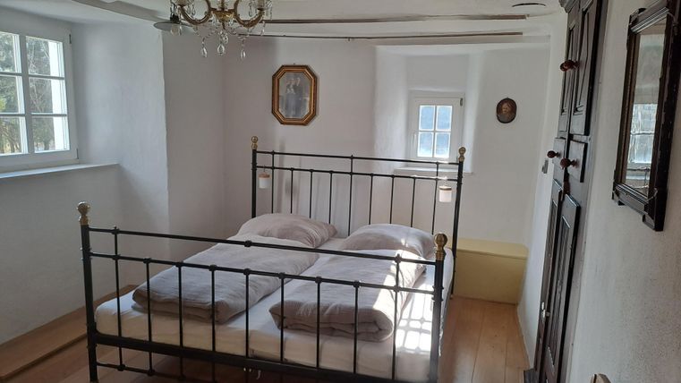 Ein gemütliches Schlafzimmer mit einem doppelten Eisenbett und beigen Bettwäsche. Es gibt ein Fenster, das viel Licht hereinlässt, und traditionelle Holzbalken an der Decke.
