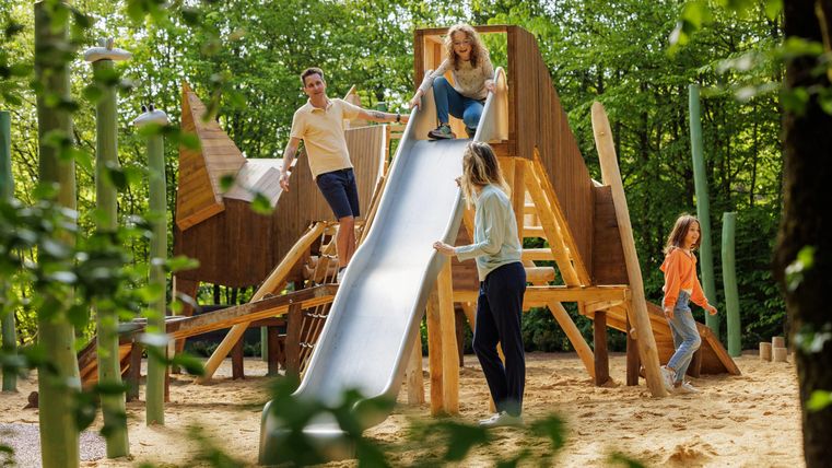 Ein Spielplatz aus Holz mit einer Rutsche, auf der Kinder spielen. Umgeben von Bäumen und einer sandigen Fläche.