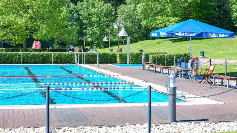 Ein Schwimmbad mit klarem Wasser, umgeben von grünen Bäumen. Am Beckenrand stehen Liegestühle und ein Sonnenschirm.