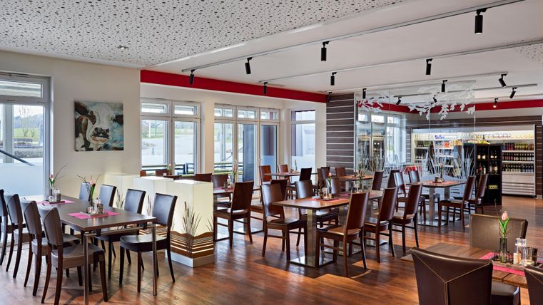 Ein modernes Restaurant mit Holzfußboden und vielen Tischen. Große Fenster sorgen für viel natürliches Licht.