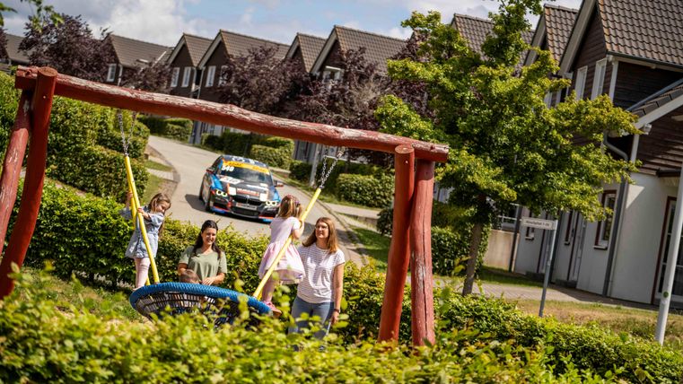 Ein Spielplatz mit Schaukeln und einem bunten Auto im Hintergrund. Einige Kinder spielen fröhlich im Freien.
