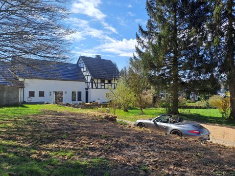 Ein Haus im ländlichen Stil mit einem großen Garten und Bäumen. Im Vordergrund steht ein silbernes Cabriolet auf einem befestigten Weg.
